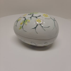 May include: White porcelain egg-shaped trinket box with a floral design. The lid features a delicate branch with white flowers and yellow centers. The bottom of the box has a simple swirl design.