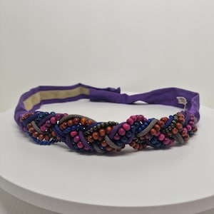 Puede incluir: Una diadema morada con un diseño trenzado con cuentas marrones, rosas, azules y grises.