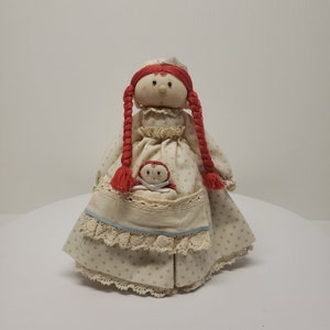 May include: A vintage handmade doll with red hair and a white dress holding a smaller doll. The doll is wearing a white dress with a blue trim and a lace trim.