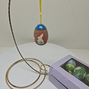 Puede incluir: Un conjunto de cuatro huevos de Pascua de madera pintados a mano en una caja púrpura con una ventana transparente. Los huevos están decorados con conejos, flores y otros motivos primaverales. Un huevo está colgado de un soporte dorado.