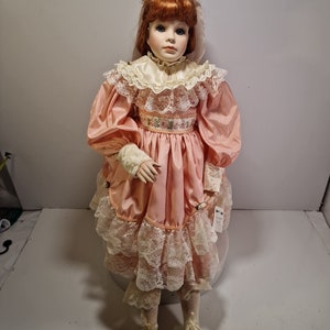 May include: A porcelain doll with red hair wearing a pink dress with lace trim and a white lace collar. The doll is wearing white shoes and has a tag on her dress.