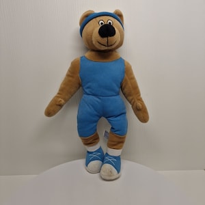 Puede incluir: Un oso de peluche marrón que lleva una diadema azul, un top azul y zapatillas azules. El oso está de pie sobre una superficie blanca.