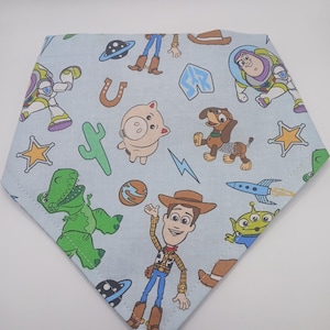 Puede incluir: Un pañuelo de color azul claro con temática de Toy Story. El pañuelo presenta personajes como Woody, Buzz Lightyear, Rex y Jamón. El pañuelo está decorado con estrellas, una herradura, un cactus y un cohete.