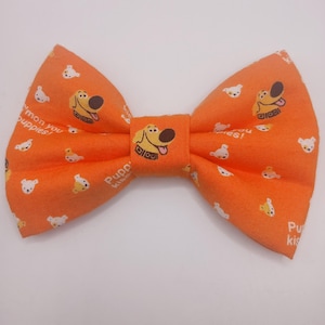Può includere: Papillon arancione con un motivo ripetuto di cani dei cartoni animati e facce di orsi bianchi. Il papillon ha il testo "Puppy Kisses" e "mon you puppies!". Un accessorio divertente per animali domestici.
