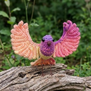 May include: A handmade crochet bird with outstretched wings, perched on a piece of driftwood. The bird features a purple head, pink and peach wings, and a yellow body. The bird's legs are made of wire. The background is blurred green foliage.