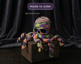 Puede incluir: Un pulpo de ganchillo hecho a mano con cabeza en forma de calavera, en tonos morado, naranja, amarillo y verde. El pulpo está sobre una caja de madera. Se ve el texto "Made to order" y "@LynnfeyCrochet".