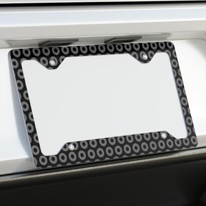 May include: A black and silver license plate frame with a circular pattern. The frame is attached to the back of a white car.