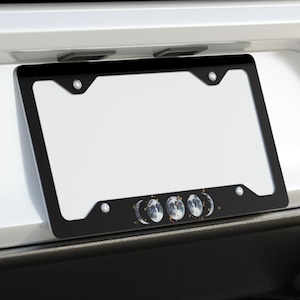 May include: A white car with a black license plate frame featuring a moon phase design. The frame has a white background and black lettering. The car has dual exhaust pipes.