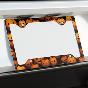 May include: A white car with a Halloween-themed license plate frame featuring a pattern of orange pumpkins. The frame is attached to the rear bumper of the car.