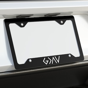 May include: A black license plate frame with a white "G>AV" logo on a white car. The frame is attached to the rear bumper of the car.