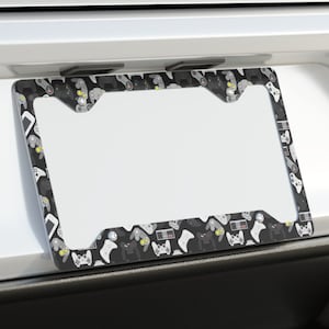 May include: A black and white license plate frame with a pattern of video game controllers. The frame is attached to a white car.