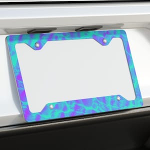 May include: A license plate frame with a vibrant teal and purple abstract design. The frame is mounted on the back of a white car, with the exhaust pipes visible below. The design has a marbled appearance.
