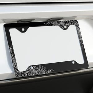 May include: A black license plate frame with a white floral design. The frame is attached to the back of a white car.