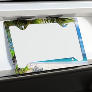 May include: A license plate frame with a tropical beach scene. The frame is silver with a colorful image of a sandy beach, palm trees, and blue sky. The frame is mounted on the back of a white car.