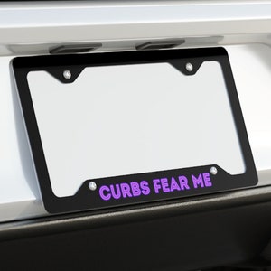 May include: Black license plate frame with the text "CURBS FEAR ME" in purple. The frame is mounted on the back of a white car. The car has dual exhaust pipes.