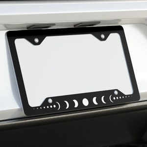 May include: A black license plate frame with a moon phase design. The frame is attached to a white car.