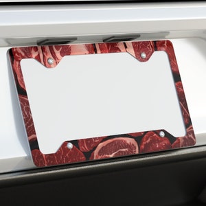 May include: A white car with a red and brown license plate frame featuring a pattern of meat. The frame is attached to the rear bumper of the car. The car has a black bumper and two chrome exhaust pipes.