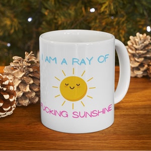May include: White ceramic mug with a yellow sun with a smiling face and the text "I am a ray of fucking sunshine" in blue and pink.