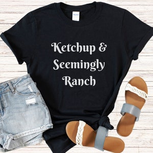 May include: Black t-shirt with white text that reads "Ketchup & Seemingly Ranch". The shirt is paired with denim shorts and sandals.