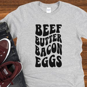 May include: A grey t-shirt with the words "BEEF BUTTER BACON EGGS" printed in black, in a retro font style.