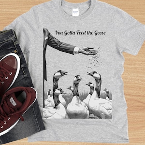 Puede incluir: Una camiseta gris con una ilustración en blanco y negro de un hombre alimentando gansos. El texto "You Gotta Feed the Geese" está impreso en la camiseta.