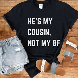 May include: Black t-shirt with white text that reads "He's my cousin, not my BF". The shirt is laid flat on a wooden surface with a pair of blue denim shorts and two pairs of sandals.