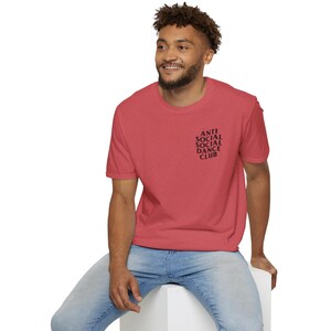 May include: A red t-shirt with the text "ANTI SOCIAL SOCIAL DANCE CLUB" printed in black on the upper left chest. The shirt is short-sleeved and has a crew neck.
