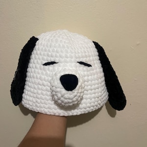 May include: A white crochet hat shaped like a dog's head, featuring black ears, a black nose, and closed eyes. The hat is made of a soft, textured yarn, and is likely a novelty item or costume accessory.