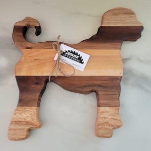 May include: A wooden cutting board shaped like a dog, with a light brown, dark brown, and walnut wood grain pattern. The board is made by ABE creations, as indicated by the tag attached to the board.