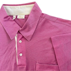 May include: A pink short-sleeved polo shirt with a white collar and a small pocket on the left side. The shirt has a button-up closure and the label "Brioni" is visible on the collar.