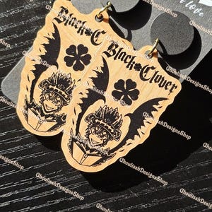 May include: A pair of wooden earrings featuring a black and white illustration of a character from the anime Black Clover. The earrings are shaped like a clover leaf with wings and the character's face is in the center. The text "Black Clover" is written on the earrings.