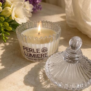 Peut inclure: Une bougie allumée dans un pot en verre transparent cannelé avec un couvercle assorti. L'étiquette indique PERLE LUMIERE FREESIA AND LILAC avec une icône de coquillage. La bougie a une mèche et de la cire blanche, près de fleurs.