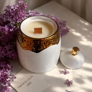 Peut inclure: Une bougie blanche avec un bord doré et une mèche en bois, accompagnée d'un couvercle assorti avec un bouton doré. La bougie est étiquetée "French Lilac" et est entourée de fleurs de lilas violettes.