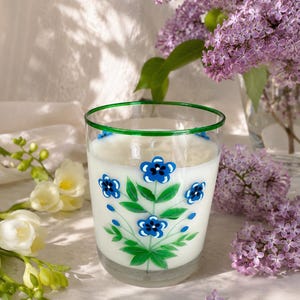 Peut inclure: Un bougeoir en verre transparent avec un bord vert, rempli de cire blanche. L'extérieur du verre présente un motif peint à la main de fleurs bleues aux centres sombres et de feuilles vertes disposées en un motif botanique vertical.