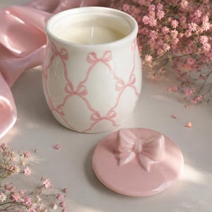 Peut inclure: Un pot à bougie en céramique blanche orné d'un motif de treillis à nœuds roses, accompagné d'un couvercle rose assorti surmonté d'un nœud sculpté. La bougie contient de la cire blanche avec une mèche centrale dans un récipient arrondi.