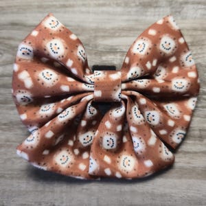 May include: A brown hair bow featuring a repeating pattern of smiling snowmen with white accents. The bow is tied in the center with a black band. The fabric has a satin sheen.