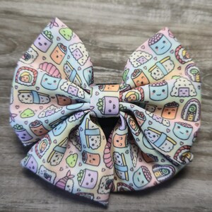 May include: A large, pastel-colored hair bow featuring a repeating pattern of cute sushi characters. The bow is tied in the center and has a variety of sushi rolls, nigiri, and other Japanese food items. The background is a soft, gradient of colors.