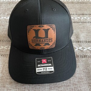 May include: Black trucker hat with a brown leather patch that says "HERNANDEZ" with a stylized letter H. The hat has a black mesh back and a white label that says "EST. 1970 RICHARDSON ORIGINAL 112 TRUCKER ADJUSTABLE".