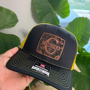 May include: Black and yellow trucker hat with a brown leather patch that says "CORONA" in a decorative font. The hat is adjustable and has a Richardson 112 label.