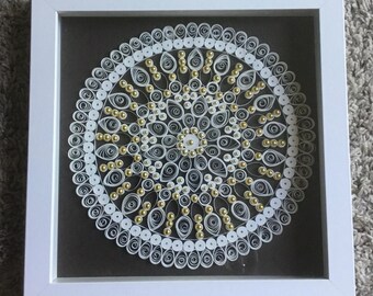 May include: A white paper mandala with gold beads in a white frame. The mandala is a circular design with intricate patterns and is surrounded by a border of gold beads.