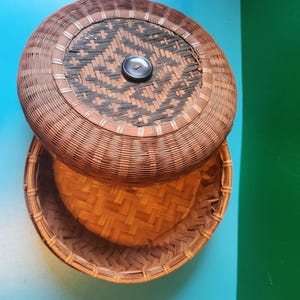 May include: A round, woven, brown basket with a lid. The lid has a woven design with a black button in the center.