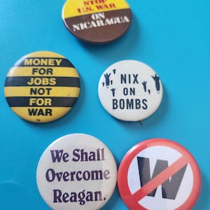 May include: A collection of five round political buttons with various slogans. The buttons feature text such as "Stop U.S. War on Nicaragua," "Money for Jobs Not for War," "Nix on Bombs," and "We Shall Overcome Reagan."