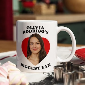 May include: White ceramic mug featuring the text "OLIVIA RODRIGO'S BIGGEST FAN" with a red heart graphic and a photo of Olivia Rodrigo. The mug has a curved handle and is on a table with a red plate and cookie.