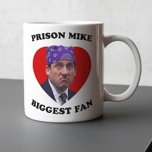 Puede incluir: Taza de cerámica blanca con el texto "PRISON MIKE BIGGEST FAN" y una imagen de un hombre con un pañuelo morado dentro de un corazón rojo. La taza tiene un asa curva y está sobre una superficie gris.