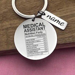May include: A silver keychain with a round tag that reads "Medical Assistant" and lists nutrition facts for an essential worker. The tag includes a list of qualities with 100% daily value, such as hard work, incredible skill, passion, intelligence, dedication, determination, and commitment. A smaller tag attached to the keychain reads "name".