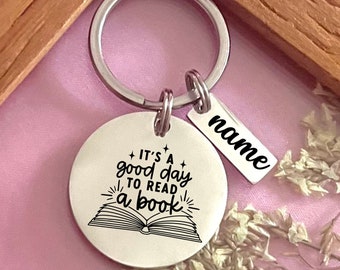 May include: Silver keychain with a round tag that says "It's a good day to read a book" with a stylized image of an open book. A smaller tag is attached to the keychain that says "name".