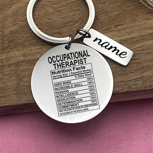 Op de afbeelding: Zilveren sleutelhanger met een rond label waarop "Occupational Therapist" staat en "Nutrition Facts" met een percentage dagelijkse waarde voor hard werken, ongelooflijke vaardigheden, passie, intelligentie, toewijding, vastberadenheid en toewijding. De sleutelhanger heeft ook een kleiner label met de tekst "name".