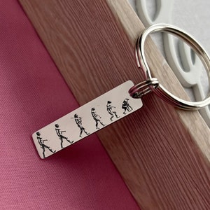 May include: A silver rectangular keychain with a series of etched figures depicting human evolution. The keychain is attached to a silver split ring. The background includes pink fabric and a wooden frame.