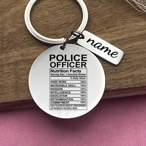 Könnte beinhalten: Ein silberner Schlüsselanhänger mit einem runden Anhänger, auf dem "Police Officer" steht und "Nutrition Facts" mit einem prozentualen Tageswert für harte Arbeit, unglaubliche Fähigkeiten, Leidenschaft, Intelligenz, Hingabe, Entschlossenheit und Engagement aufgelistet sind. Der Anhänger enthält auch den Text "Serving Size: 1 Essential Worker". Ein kleinerer silberner Anhänger mit dem Wort "name" hängt am Schlüsselanhänger.