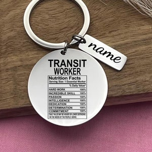 Könnte beinhalten: Silberner Schlüsselanhänger mit einem runden Anhänger, auf dem "Transit Worker" steht und "Nutrition Facts" mit einem prozentualen Tageswert für harte Arbeit, unglaubliche Fähigkeiten, Leidenschaft, Intelligenz, Hingabe, Entschlossenheit und Engagement aufgelistet sind. Der Anhänger enthält auch den Text "Serving Size: 1 Essential Worker". Ein kleinerer silberner Anhänger mit dem Wort "name" ist am Schlüsselanhänger befestigt.